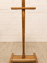 Load image into Gallery viewer, A vintage wooden coat rack with a warm medium-brown finish, featuring a central post, a horizontal bar, and a slanted base, set against a white brick wall and beige rug.
