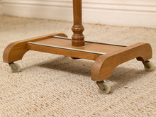 Load image into Gallery viewer, A close-up of a wooden rolling stand or table base with curved legs, a top shelf, and small caster wheels, shown on a beige carpeted floor.
