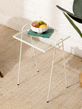 Load image into Gallery viewer, A small white metal side table with a clear glass top, styled with a bowl of fruit and a book on a beige woven rug against a white brick wall.
