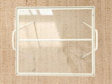 Load image into Gallery viewer, A square white-framed glass window or door panel with integrated side handles, shown from above on a textured beige woven surface.
