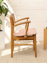 Load image into Gallery viewer, A mid-century modern wooden dining chair with a light pink cushioned seat and backrest, set against a white brick wall with natural sunlight and shadows.
