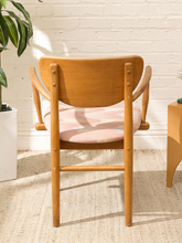 Load image into Gallery viewer, A mid-century modern wooden dining chair with a curved backrest, armrests, and a light pink cushion, set against a white brick wall with a potted plant on the left.
