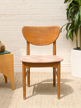 Load image into Gallery viewer, A mid-century modern wooden dining chair with a curved backrest and a light pink upholstered seat, set against a white brick wall with a potted plant and side table.
