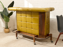 Load image into Gallery viewer, A mid-century modern rolling bar stool with mustard yellow tufted leather upholstery and a dark wood frame, set in a room with a white brick wall, a potted plant, and a black chair partially visible.
