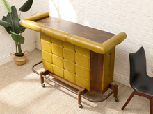 Load image into Gallery viewer, A mid-century modern bar cart with mustard yellow tufted leather upholstery, a dark wood top, and a metal rolling frame, shown in a styled room corner next to a black chair and a potted plant.
