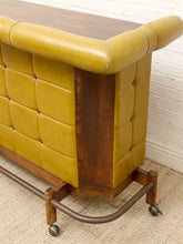 Load image into Gallery viewer, A close-up of a mid-century modern console table featuring mustard yellow tufted leather upholstery, dark brown wood paneling, and a dark metal wheeled base, set against a white brick wall and beige carpet.
