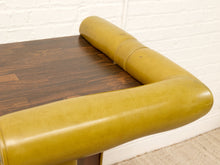 Load image into Gallery viewer, A close-up of a mustard yellow leather upholstered table corner with a dark wood tabletop, set against a white brick wall and light carpet.
