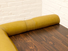 Load image into Gallery viewer, A close-up of a mustard yellow upholstered bar stool or booth cushion resting on a dark wood table, with a white painted brick wall in the background.
