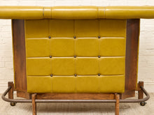 Load image into Gallery viewer, A mid-century modern bar cart featuring a mustard yellow tufted leather front panel and a dark brown wood frame with a bamboo-style top and metal rolling legs.
