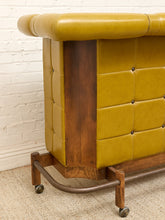 Load image into Gallery viewer, A close-up of a mid-century modern console table featuring mustard yellow tufted leather upholstery and a dark brown wood frame with a curved metal base on casters, set against a white brick wall and beige carpet.

