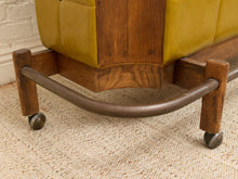 Load image into Gallery viewer, Close-up of a mid-century modern rolling ottoman or stool with mustard yellow leather upholstery, a dark wood frame, and a curved metal base on casters, set on a beige woven rug.
