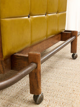 Load image into Gallery viewer, Close-up of the lower portion of a vintage upholstered rolling chair or sofa, showing mustard yellow tufted leather, a dark wood frame, and a metal tube base with casters on a beige carpet.
