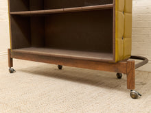 Load image into Gallery viewer, A mid-century modern rolling bookcase with a dark wood frame, a tufted mustard yellow leather side panel, and a three-tier shelf, shown on a beige carpet against a white brick wall.

