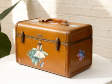 Load image into Gallery viewer, A vintage brown leather trunk with floral and bird illustrations, sitting on a white surface against a white brick wall.
