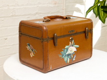 Load image into Gallery viewer, A vintage brown leather suitcase or trunk with brass latches and handle, decorated with painted bird and floral designs, sitting on a white table against a white brick wall.
