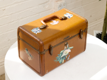 Load image into Gallery viewer, A vintage brown leatherette jewelry box with painted floral and bird illustrations, resting on a white round table against a white brick wall.
