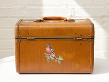Load image into Gallery viewer, A vintage brown leather suitcase with brass hardware and a painted bird on the front, set against a white brick wall background.
