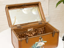 Load image into Gallery viewer, A vintage brown leather trunk with an open glass lid, showing a satin lining and floral painted details on the front, set against a white brick wall background.
