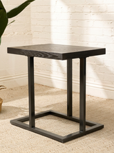 Load image into Gallery viewer, A square black end table with a dark wood-grain top and a geometric metal frame base, set on a beige carpet against a white brick wall.
