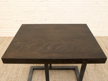 Load image into Gallery viewer, A square dark brown wood table with a thick top and a black metal frame base, set against a white brick wall and light woven rug.
