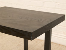 Load image into Gallery viewer, Close-up of a dark wood table top with a black finish and visible grain, supported by black metal legs against a white brick wall and light floor.
