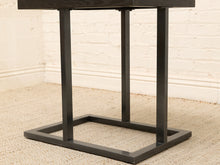Load image into Gallery viewer, A close-up of a dark gray or black metal table base with a square frame and three vertical legs, set on a beige textured carpet against a white brick wall.
