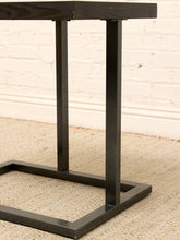 Load image into Gallery viewer, A close-up of a black metal side table with a rectangular top, two vertical legs, and a square base, set on a woven rug against a white brick wall.
