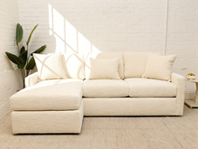 Load image into Gallery viewer, A cream-colored upholstered sectional sofa with a left-facing chaise lounge, styled in a bright room with a white brick wall, a potted plant, and a small side table.
