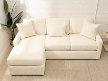 Load image into Gallery viewer, An off-white upholstered sectional sofa with a left-facing chaise lounge and four matching throw pillows, set in a bright room with a white brick wall, beige rug, and a potted plant.
