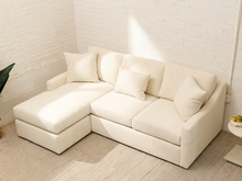 Load image into Gallery viewer, A cream-colored modular sectional sofa with a chaise lounge, styled in a bright, minimalist room with white brick walls and a light-colored rug.
