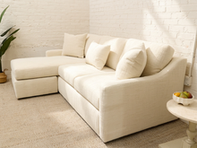Load image into Gallery viewer, A cream-colored upholstered sectional sofa with a chaise lounge on the left, styled in a bright, modern room with a white brick wall, beige rug, and a small side table.
