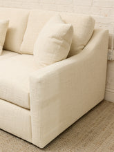 Load image into Gallery viewer, Close-up of a cream-colored upholstered sofa corner with textured fabric, matching throw pillow, woven rug, and a white brick wall background.

