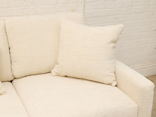 Load image into Gallery viewer, Cream-colored textured fabric sofa with matching throw pillows, set against a white brick wall background.
