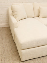 Load image into Gallery viewer, A cream-colored upholstered sectional sofa with textured fabric, featuring a chaise lounge and matching throw pillows, set against a white brick wall and a woven jute rug.
