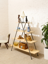 Load image into Gallery viewer, A four-tier A-frame wooden shelf with dark metal legs, styled with various decorative objects including vases, sculptures, and a small trunk, set against a white brick wall.
