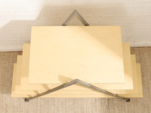 Load image into Gallery viewer, A stack of light natural wood rectangular boards or panels displayed on a metal V-shaped stand, photographed from above against a white brick wall and woven rug.

