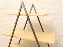Load image into Gallery viewer, A modern three-tier A-frame shelving unit with light natural wood shelves and a dark gray metal frame, set against a white brick wall.
