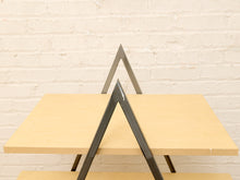 Load image into Gallery viewer, A close-up of a modern table with light natural wood square tops and dark charcoal gray triangular metal legs, set against a white painted brick wall.
