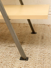 Load image into Gallery viewer, Close-up of a light wood tabletop and industrial metal legs with square black base plates, resting on a textured beige woven rug.
