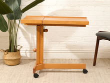 Load image into Gallery viewer, A light natural wood folding table with black caster wheels, shown in a styled room setting with a white brick wall, a potted plant, and a chair leg visible.
