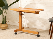 Load image into Gallery viewer, A light natural wood lecture table with a height-adjustable central column, black caster wheels, and a front storage tray, shown in a room setting against a white brick wall.
