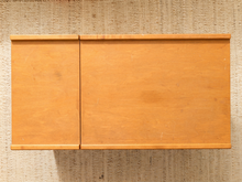 Load image into Gallery viewer, A light natural wood cabinet or wall-mounted shelf with two doors, shown against a textured beige woven wall.
