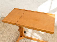 Load image into Gallery viewer, A light brown wooden folding table with a fold-down top section, supported by a T-style base with casters, set on a beige carpet against a white brick wall.
