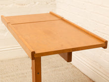 Load image into Gallery viewer, A light brown wooden folding table with a slatted top, shown in a room setting against a white brick wall and beige carpet.
