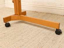 Load image into Gallery viewer, Close-up of a wooden rolling stand or easel base with black caster wheels on a beige textured carpet in front of a white wall and baseboard.
