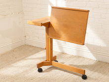 Load image into Gallery viewer, A light natural wood mobile writing desk with a fold-down screen and small side tray, mounted on a wheeled base, set against a white brick wall and beige carpet.
