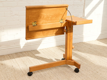 Load image into Gallery viewer, A light natural wood lecture podium with a hinged back panel, small side table, and caster wheels, shown in a bright room with a white brick wall and beige carpet.
