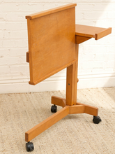 Load image into Gallery viewer, A light wood rolling lecture desk with a fold-down writing surface, side shelf, and three-star wheeled base, photographed against a white brick wall and beige carpet.

