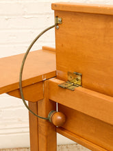 Load image into Gallery viewer, Close-up of a light orange-brown wooden table or desk showing the hinge mechanism, brass hardware, and a round wooden pull knob connected by a curved metal cable.
