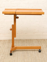 Load image into Gallery viewer, A mid-century modern wooden trolley table with a light oak finish, featuring a fold-down writing surface, adjustable height knobs, and black caster wheels, set against a white brick wall and beige rug.

